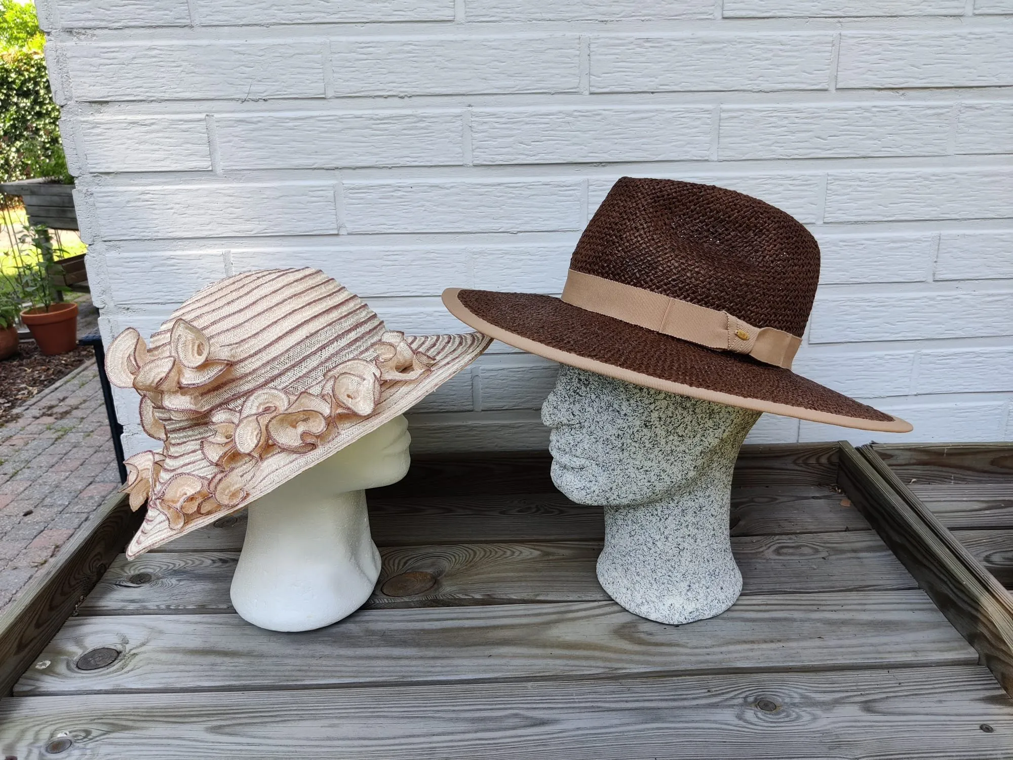 Roomkleurige hoed met organza bloemen langs de rand, naast een bruine fedora in grof stro
