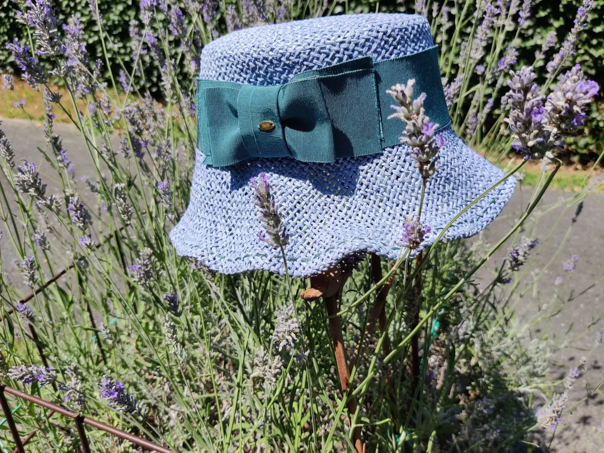 Lichtblauwe gevlochten buckethoed met donkergroene grosgrain strik, tussen de lavendel