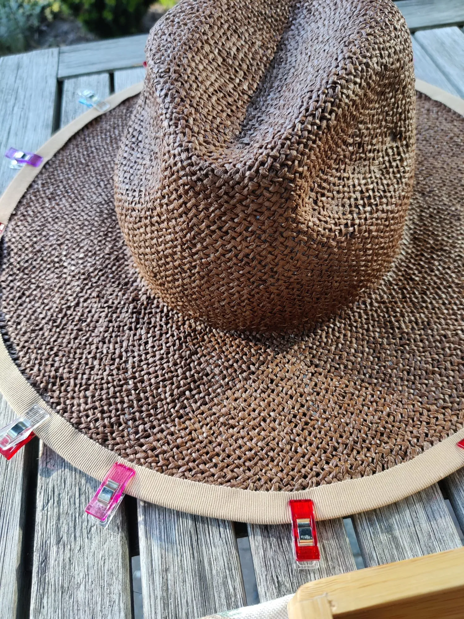 Brede bruine strohoed op tafel, met naaiklemmen die het beige lint langs de rand op zijn plaats houden